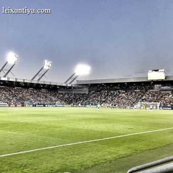 A large soccer stadium filled with spectators under bright floodlights. The field is green and well-maintained with players actively engaged in a match. Advertising banners line the perimeter of the stadium. A cameraman is positioned near the foreground, capturing the action on the field. The sky above is clear with a hint of dusk.