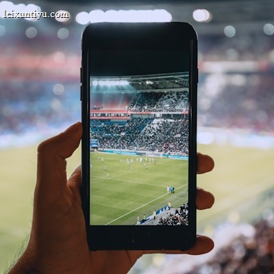 A hand holding a smartphone captures a live view of a soccer stadium filled with fans and players on the field. The image on the smartphone screen is crisp, showing a wide-angle view of the game in action.