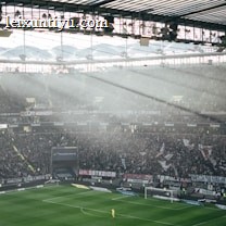 A large football stadium filled with spectators during a match. Sunlight filters through the transparent roof, illuminating parts of the stands and the field. A digital scoreboard displays a score of 1 to 0 at 46 minutes and 37 seconds. Banners and flags are visible among the dense crowd, adding to the vibrant atmosphere.