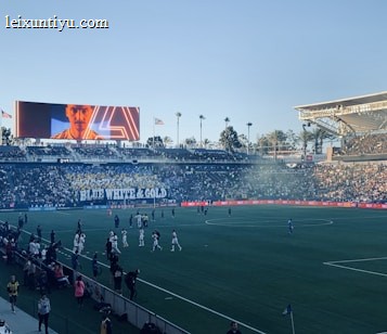 A large sports stadium filled with spectators. The field is occupied by soccer players, some walking and others scattered across the pitch. A massive screen displays a person, and flags fly on top of the stadium roof. The audience is dense, and the atmosphere seems lively with a visible banner showing 'BLUE WHITE & GOLD'.