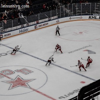 A professional ice hockey match captured from a high vantage point, featuring players in red and white uniforms moving across the rink. The scene includes a large digital screen displaying a live feed of the game and a sparsely filled audience watching from the stands.