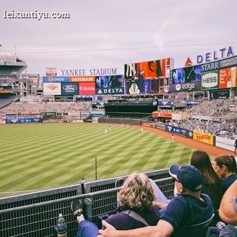A baseball stadium filled with spectators watching a game on a neatly maintained grass field. The stadium features large advertising screens displaying various logos and brands. Fans sitting in the foreground are dressed casually, enjoying the sporting event.