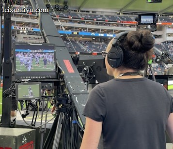 A person with headphones is operating a large camera setup at a sports stadium. The camera equipment is focused on a screen showing a live sports event taking place on a football field. The stadium is filled with spectators, and the atmosphere is busy with technical setup and monitors.