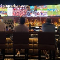 A group of people sit at a bar with their backs to the camera, watching multiple sports games displayed on large screens. The bar counter is lined with various bottles of liquor, and the atmosphere suggests a casual and relaxed social setting.